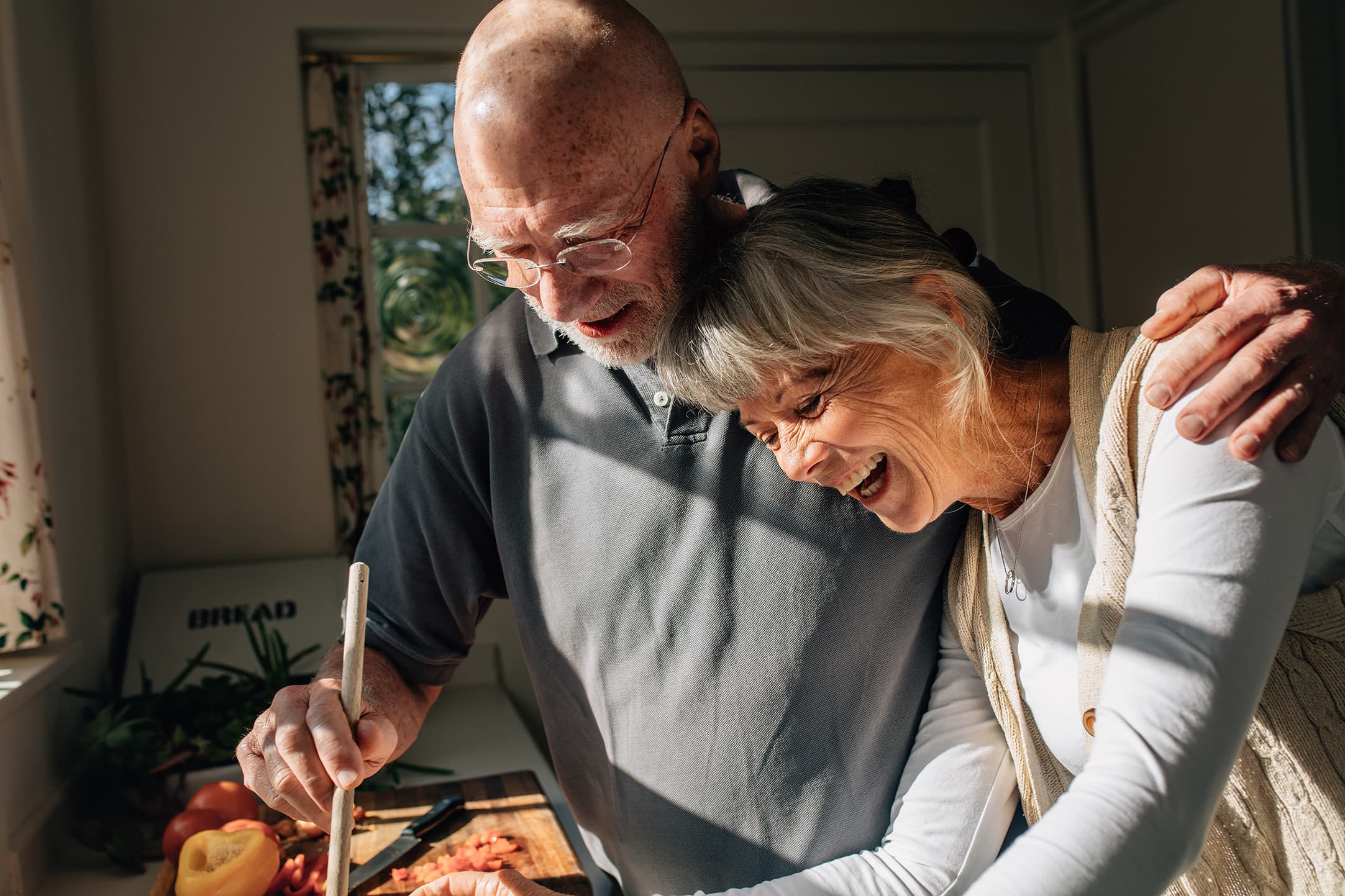 Smiling older couple preparing food together in a sunlit kitchen, capturing shared activity, joy, and healthy aging at home.